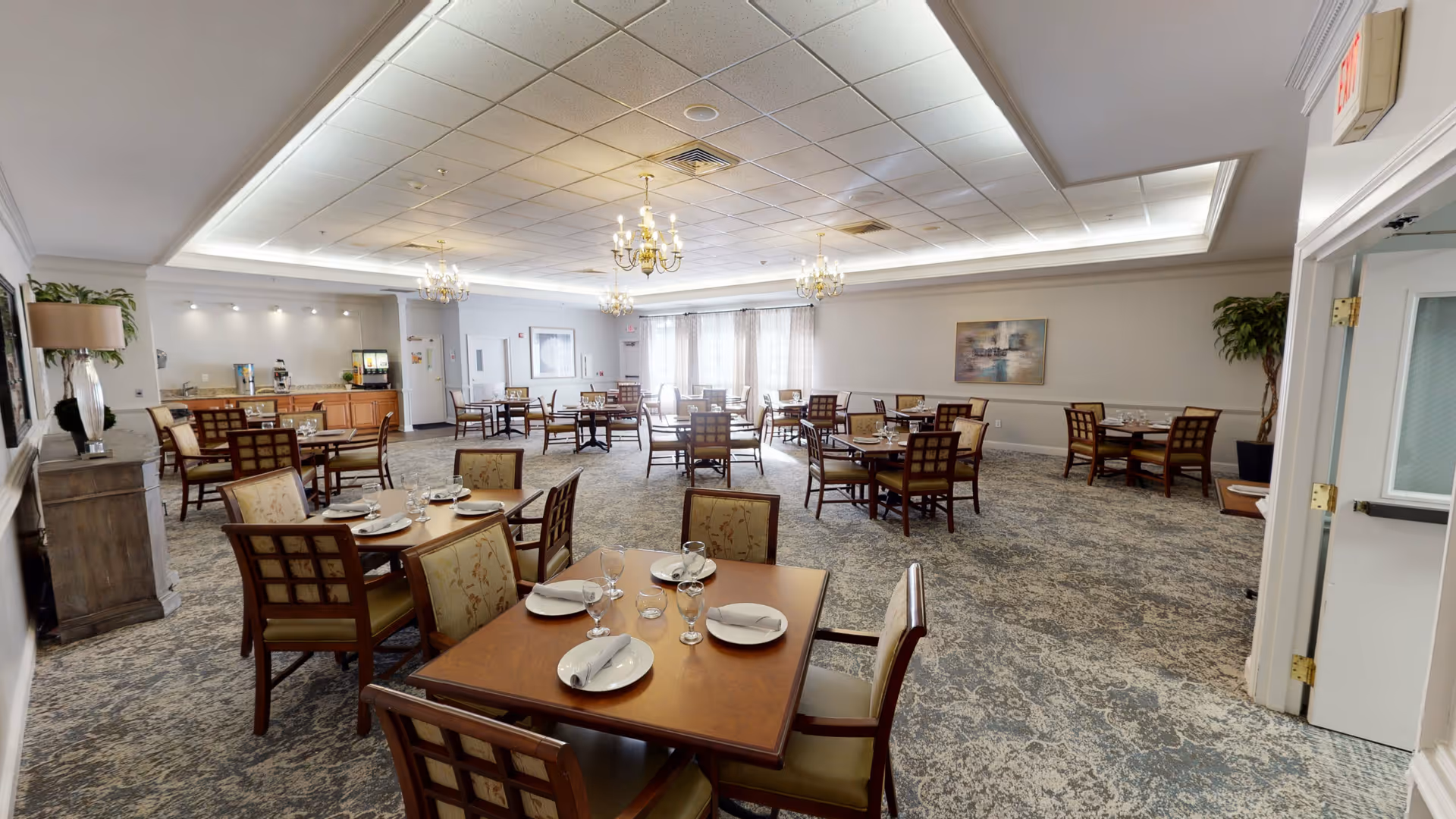 A spacious dining room with multiple wooden tables and cushioned chairs arranged neatly. Each table is set with white plates, napkins, and glassware. The room features a patterned carpet, chandeliers hanging from a drop ceiling, and large windows with sheer curtains allowing natural light to enter. There are plants and framed artwork on the walls, and a beverage station with dispensers is visible in the background.