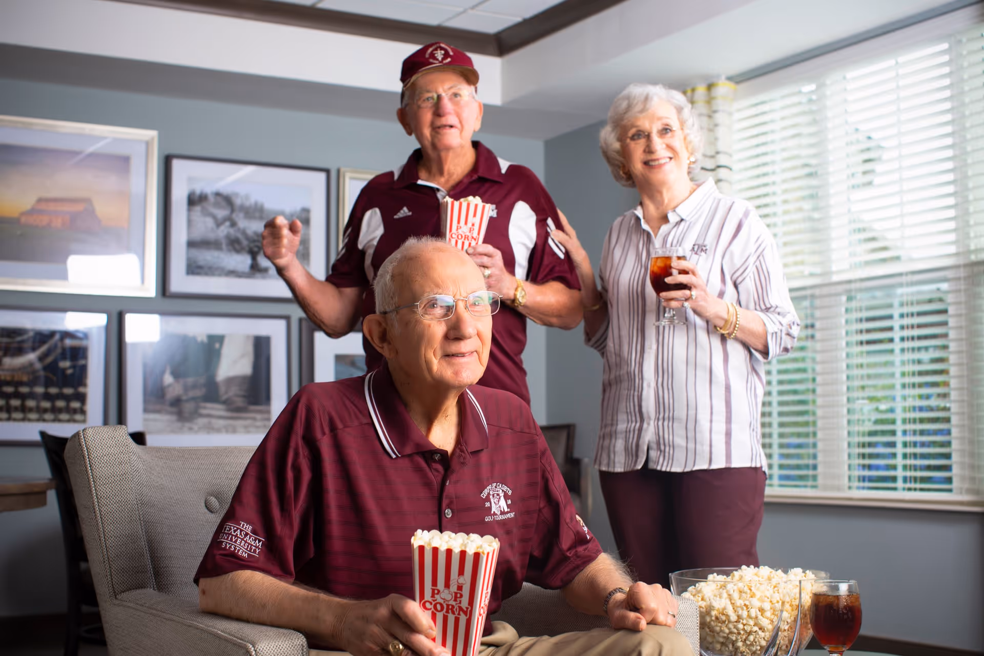 Three elderly people enjoying snacks and drinks in a cozy room. One man is seated holding a container of popcorn, while another man and a woman stand behind him, also holding popcorn and a drink respectively. The room has framed pictures on the wall and a window with blinds letting in natural light.