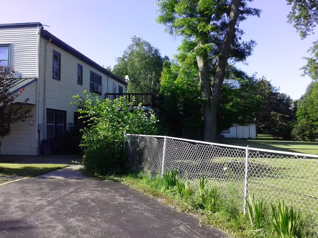 Exterior view of a two-story light-colored house with a chain-link fence, driveway, deck, and surrounding trees and lawn.