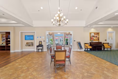 A spacious and well-lit common area in a senior living facility featuring a central dining table with chairs, a chandelier hanging from the ceiling, a piano with two yellow chairs on the right, and an open doorway leading to an outdoor patio with red umbrellas and greenery.
