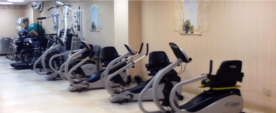 A room with several exercise machines including recumbent bikes and other fitness equipment lined up against a beige wall with two small decorative windows and a plant. There are also wheelchairs and walkers visible in the background.