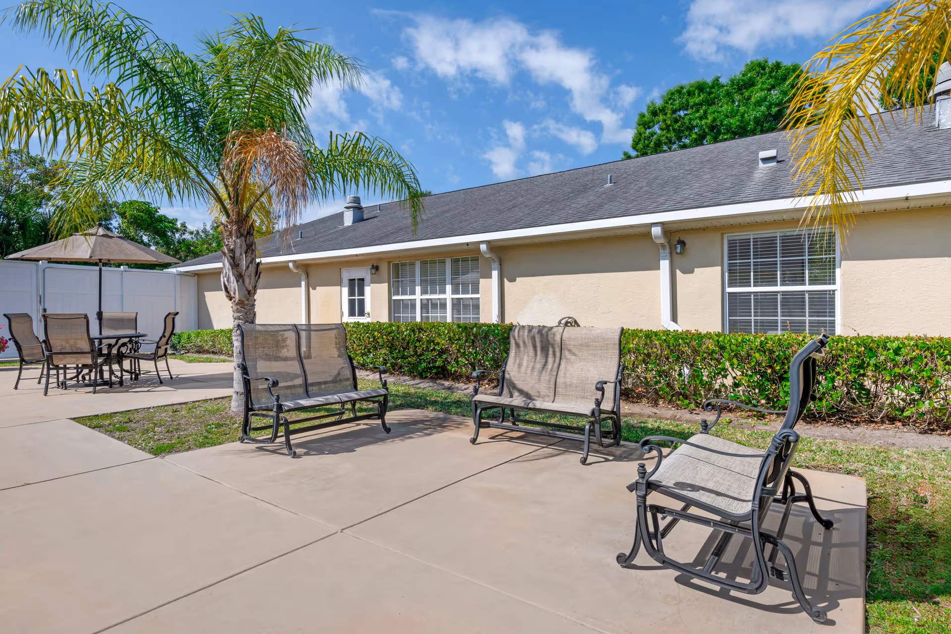 Outdoor patio area with metal benches and chairs, palm trees, and a single-story building under a blue sky.