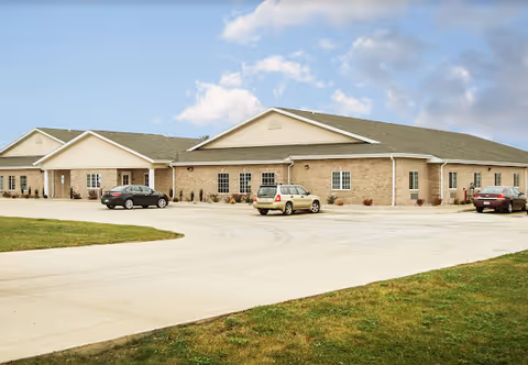 Exterior view of a single-story brick building with a green roof, several windows, and a paved parking area with three parked cars under a partly cloudy sky.