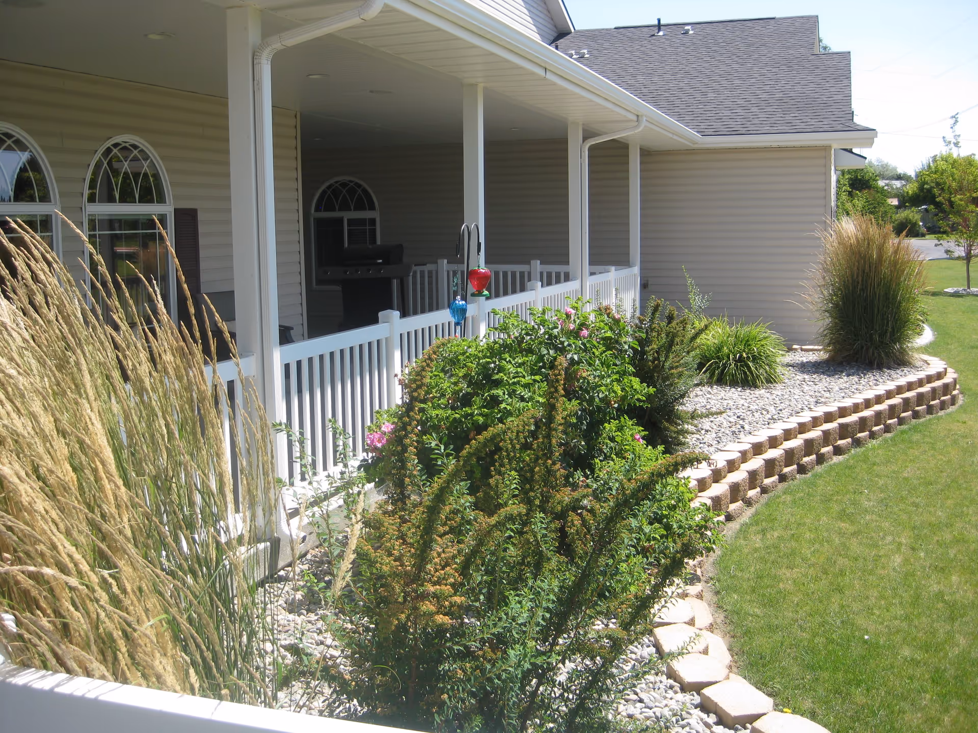 Outdoor view of a garden area next to a beige building with a covered porch. The garden features various green shrubs, ornamental grasses, and flowering plants bordered by stone edging. The porch has white railings and columns, with a barbecue grill visible inside.