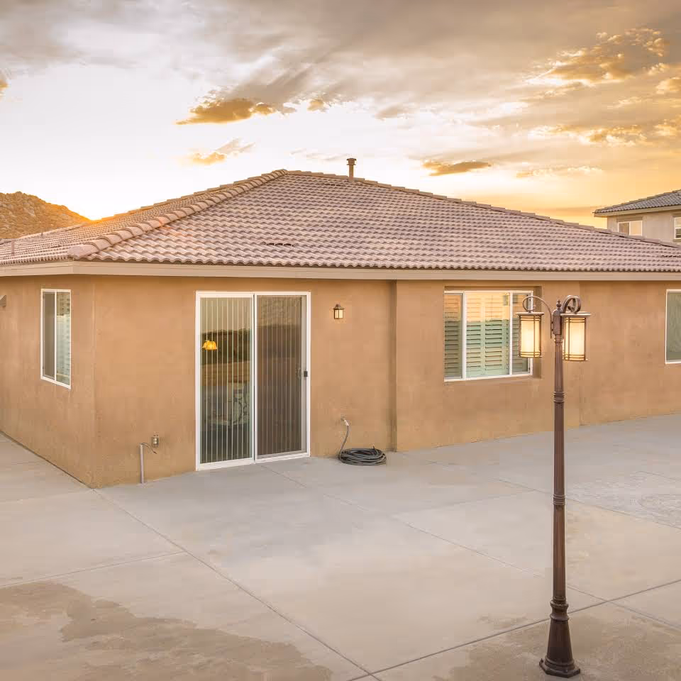 Exterior view of a single-story residential building with a tiled roof, beige stucco walls, sliding glass door, and windows. A vintage-style street lamp is visible on a concrete patio area during sunset.