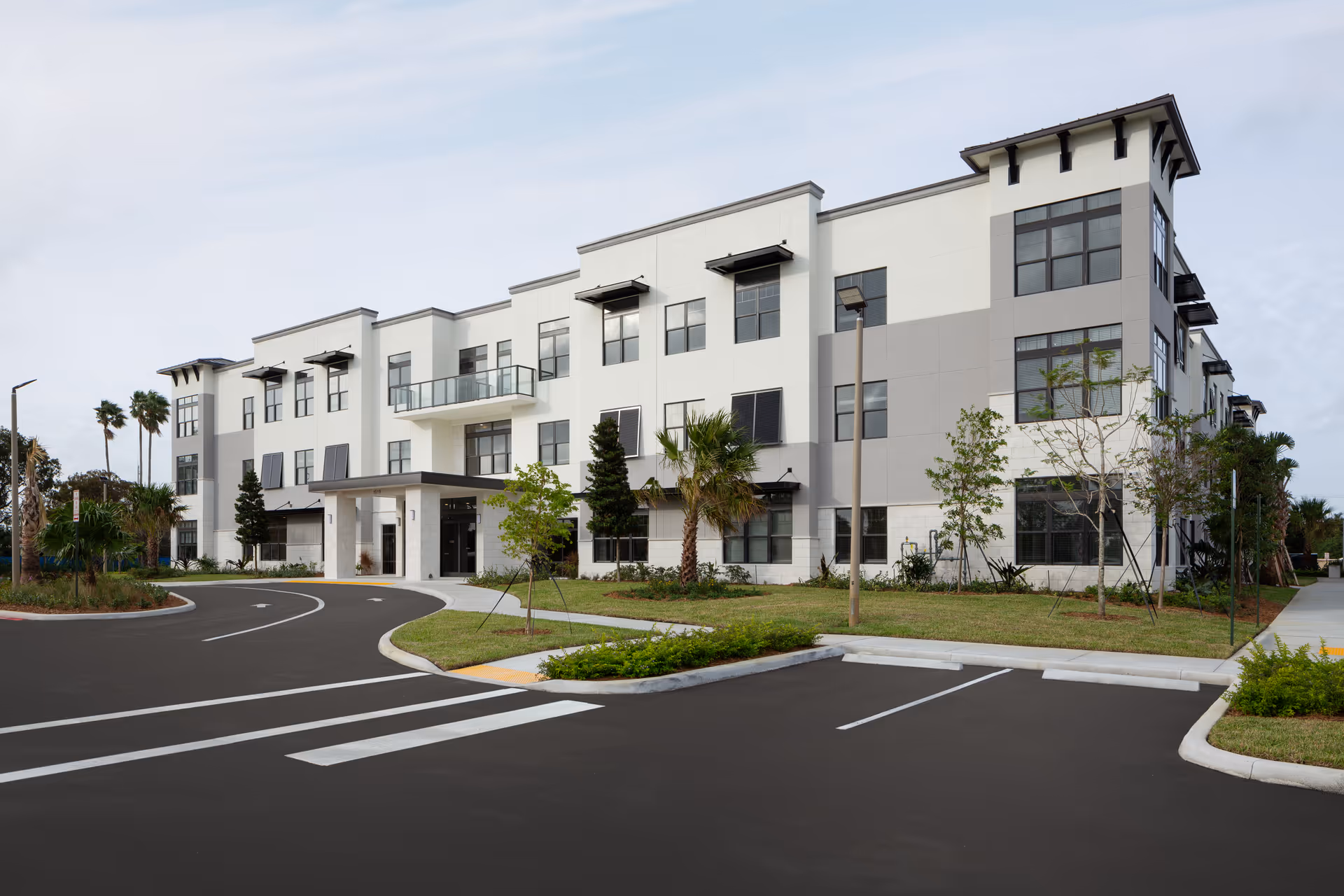 Exterior view of a modern three-story senior living facility building with large windows, a covered entrance, landscaped greenery, palm trees, and an empty parking lot in front under a cloudy sky.