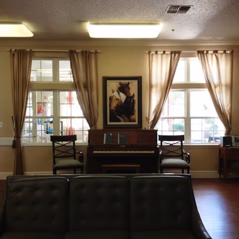 Interior view of a living room area with a dark leather sofa in the foreground, two chairs flanking an upright piano against a wall with two windows covered by beige curtains, and a framed painting of horses above the piano.