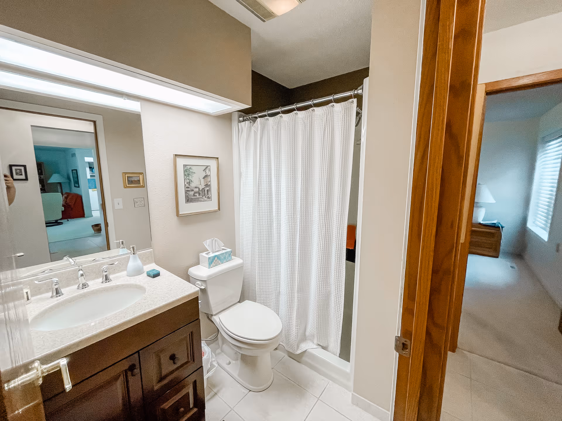 A clean bathroom with a white toilet, a sink with a marble countertop and dark wooden cabinet, a large mirror above the sink, and a shower with a white curtain. The bathroom has beige walls and tiled floor. A framed picture hangs on the wall above the toilet. The bathroom door is open, showing a glimpse of an adjacent room with a window and a lamp on a wooden nightstand.