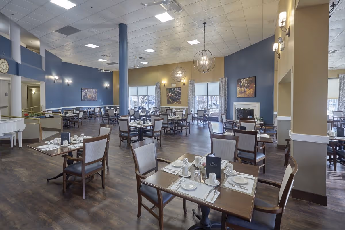 A spacious dining room in a senior living facility with multiple tables set with white plates, cups, and silverware. The room features wooden floors, blue and beige walls, large windows with curtains, modern pendant lights, and framed artwork on the walls. There is also a white piano in the corner.