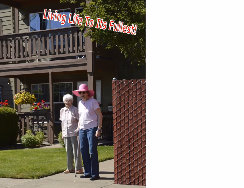 Two elderly women standing outside near a residential building with a wooden balcony and flower pots. One woman is wearing a pink hat and using a cane, while the other is smiling and standing beside her. There is a red fence and green lawn in the background. Text above reads 'Living Life To Its Fullest!'