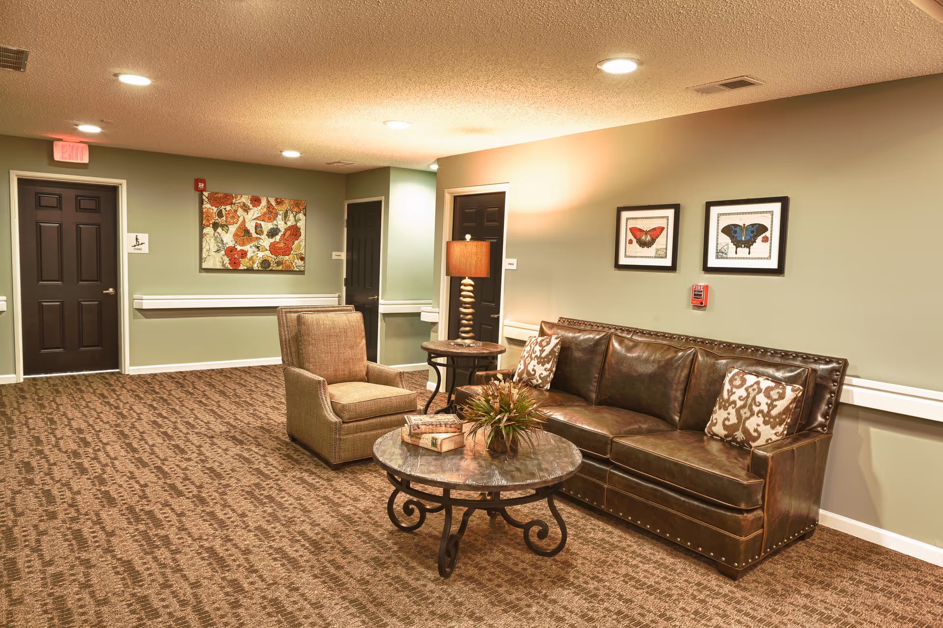 A cozy seating area in a senior living facility hallway featuring a brown leather sofa with patterned pillows, a cushioned armchair, a round coffee table with books and a plant, and a side table with a lamp. The walls are painted light green and decorated with framed butterfly artwork and a floral painting. There are three dark brown doors and a carpeted floor with a patterned design.