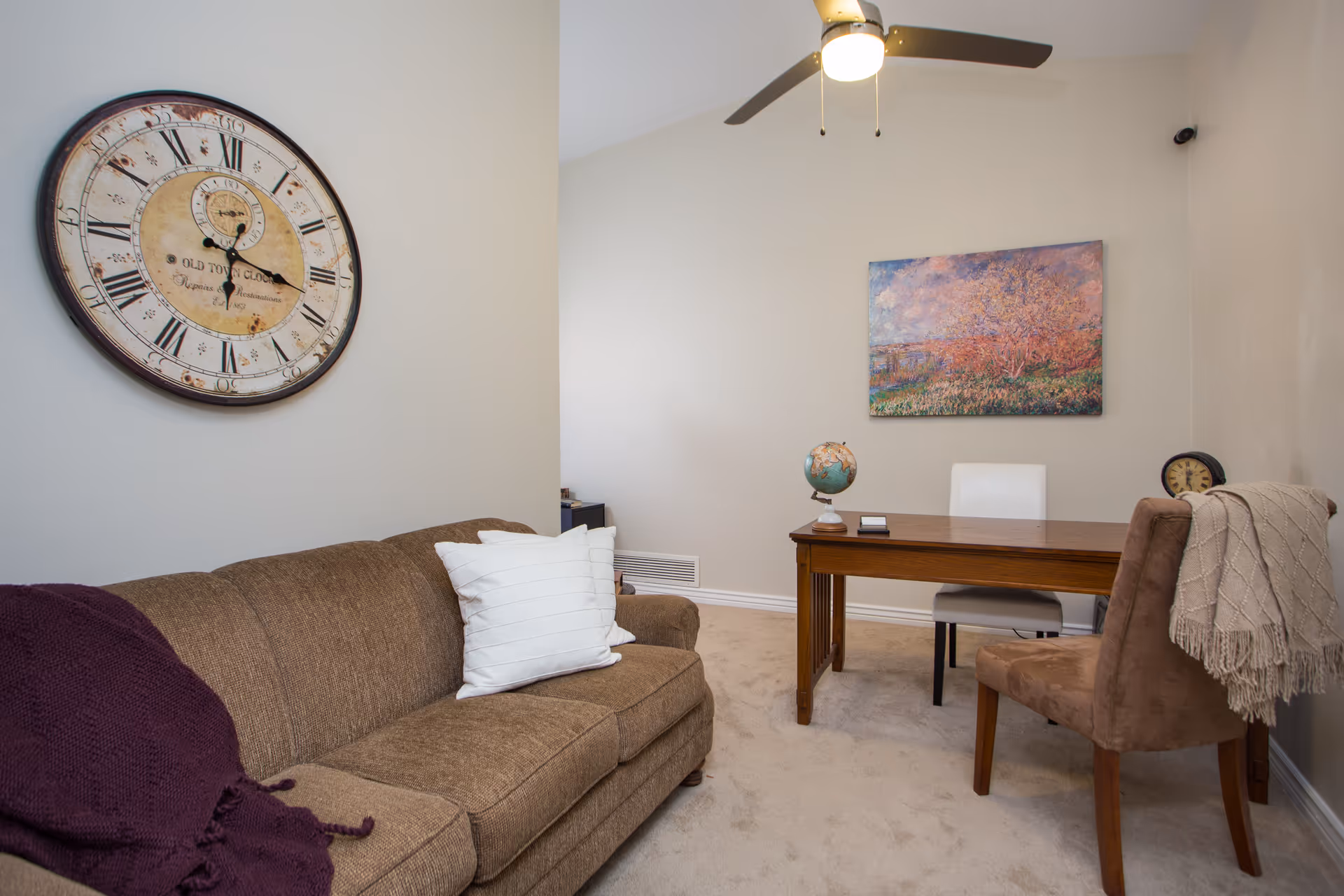 A cozy office or small sitting room with a brown sofa adorned with white pillows and a purple throw blanket. There is a wooden desk with a globe and a white chair behind it, and a brown chair with a beige throw draped over the back. A large vintage-style wall clock hangs on the left wall, and a colorful landscape painting is on the wall behind the desk. The room has beige carpet and a ceiling fan with a light.