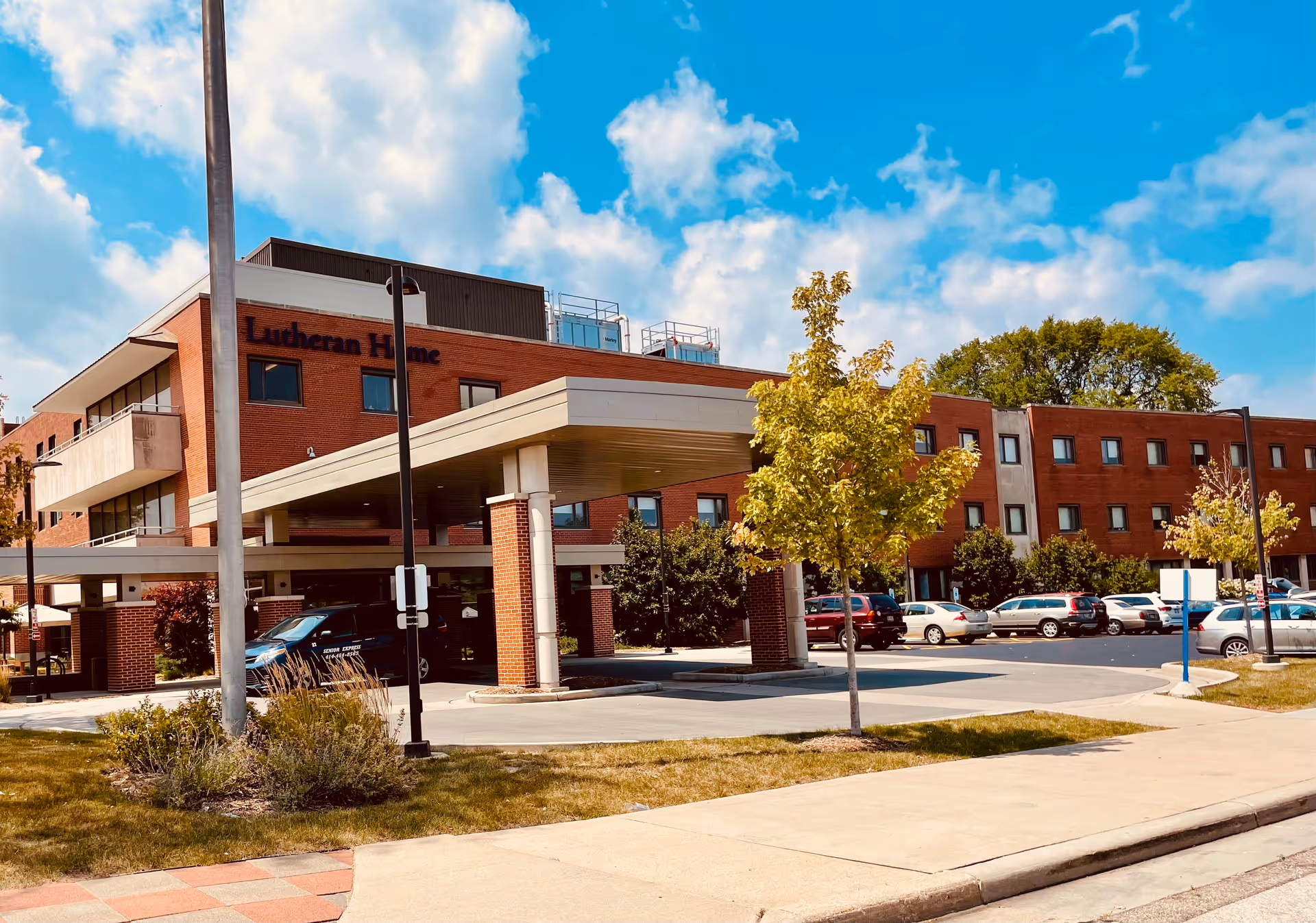 Front exterior of a brick multi-story senior living building labeled "Lutheran Home" with a covered entrance, parked cars, and trees under a blue sky.