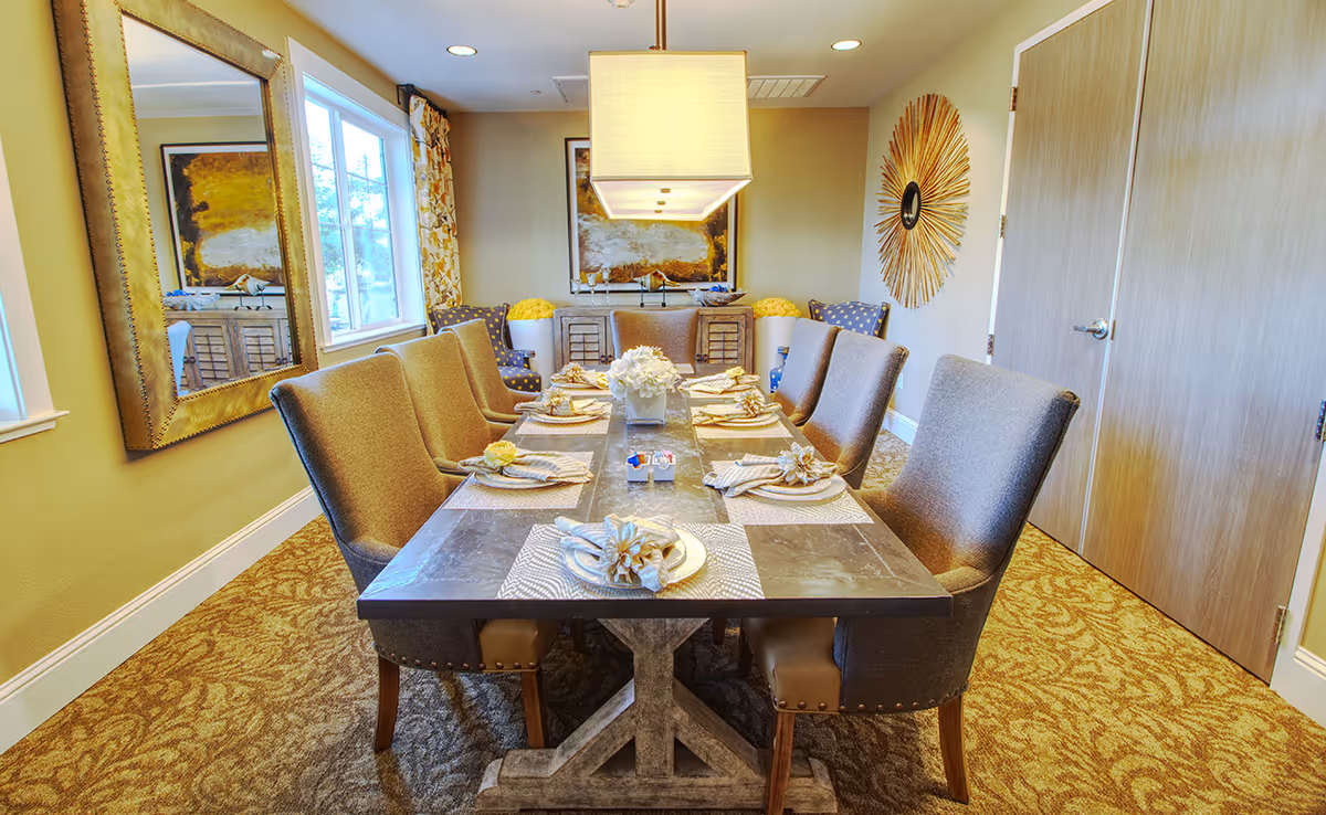 A well-lit dining room with a rectangular dining table set for eight people. The table has a dark stone top and is decorated with placemats, napkins with flower napkin rings, and a centerpiece of white flowers. Surrounding the table are cushioned chairs in a neutral tone. The room features a large mirror on one wall, a window with patterned curtains, a sideboard with decorative items, and a sunburst wall decoration. The carpet has a patterned design in warm tones.