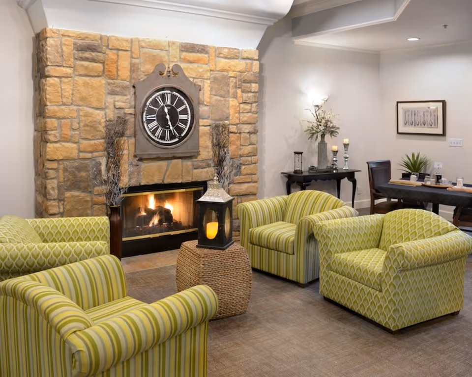 A cozy living room area with four green patterned armchairs arranged around a woven ottoman with a lantern on top. Behind the seating area is a stone fireplace with a large clock mounted above it. To the right, there is a small black table with decorative items and a dining table with chairs against a white wall.