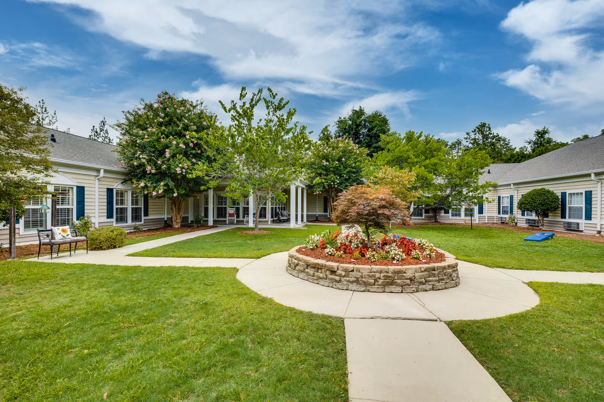 A well-maintained outdoor courtyard area at The Legacy of Camden featuring a circular flower bed with colorful flowers and a small tree in the center, surrounded by green grass and paved walkways. The courtyard is enclosed by single-story buildings with beige siding, blue shutters, and multiple windows. There are benches and trees around the area under a partly cloudy sky.