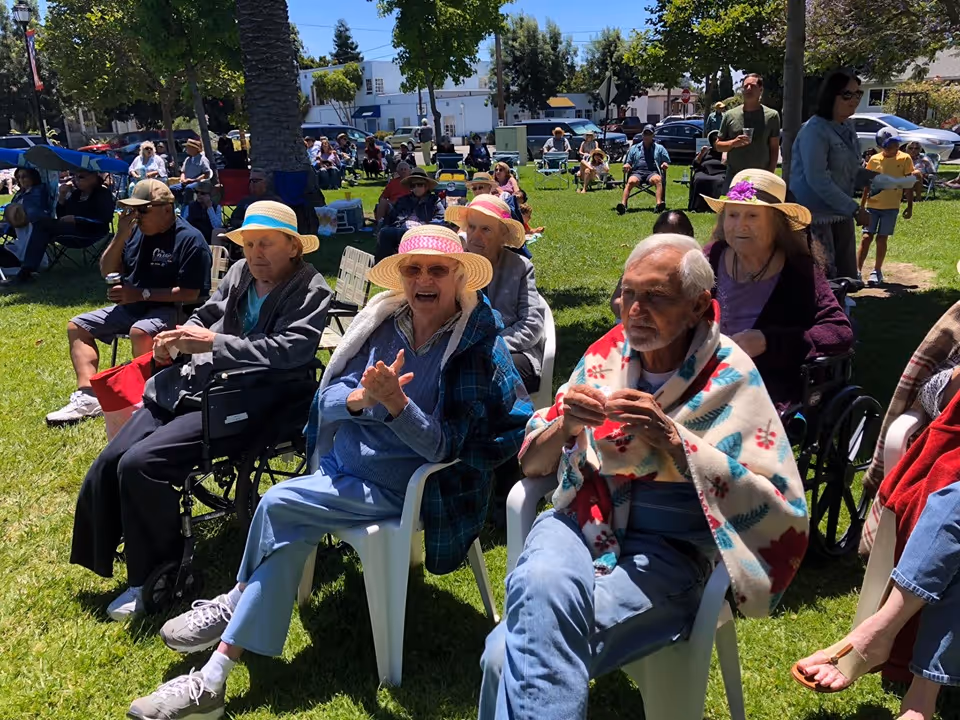 A group of elderly people sitting outdoors on a sunny day, some in wheelchairs and others in plastic chairs, wearing hats and light jackets or blankets. They are gathered on a grassy area with trees and buildings in the background, enjoying an event or gathering.