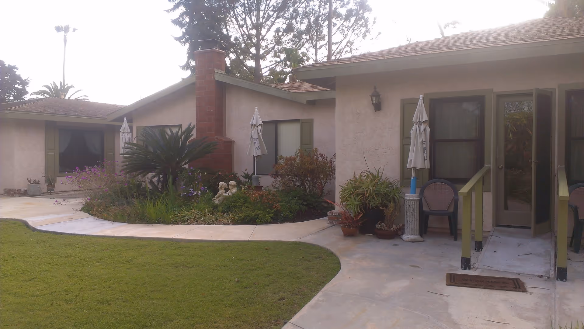 Exterior view of a single-story residential building with beige walls and green window shutters. There is a small garden with various plants and flowers, a brick chimney, and a concrete pathway leading to the entrance. Two closed patio umbrellas and plastic chairs are visible near the door.