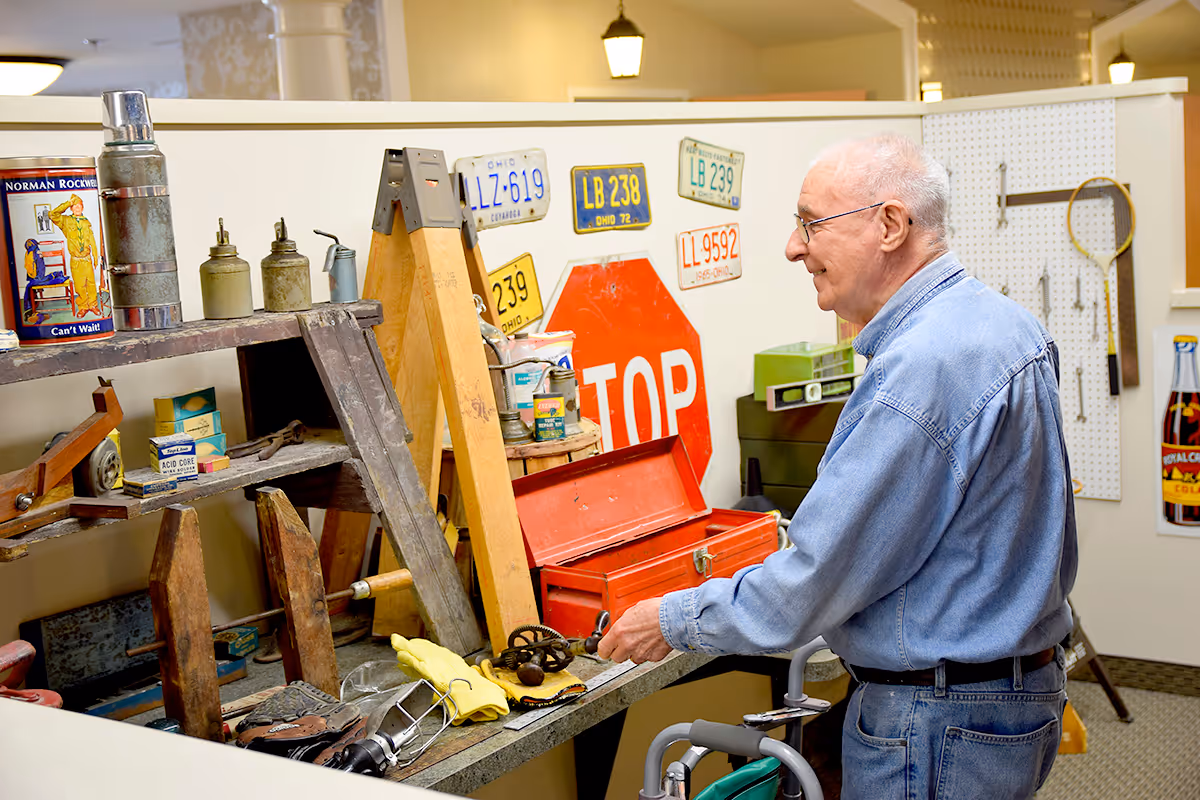An elderly man wearing glasses and a denim shirt stands with a walker in front of a display of vintage tools, old license plates, a red stop sign, and various antique items in a well-lit indoor setting.