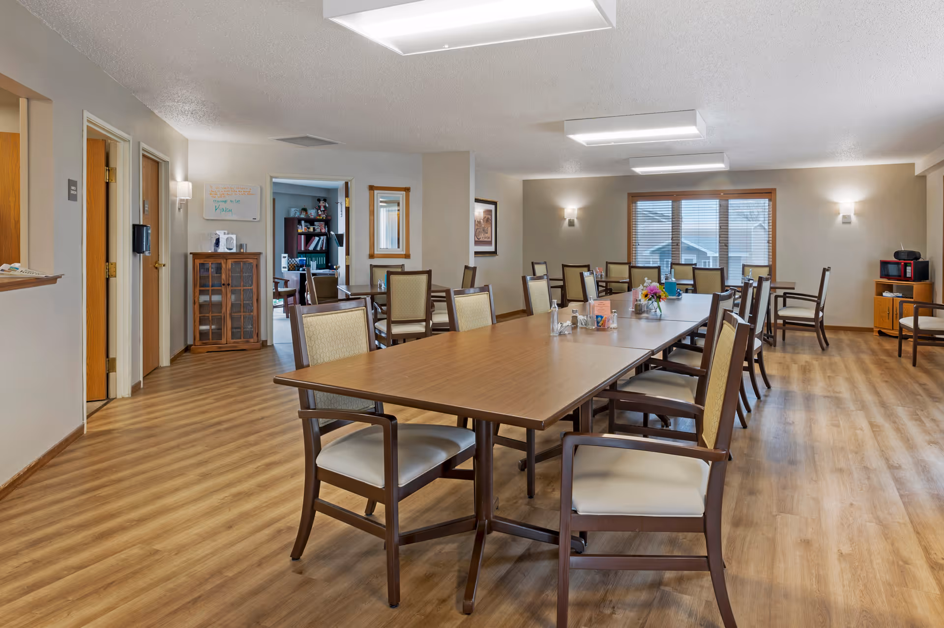 A spacious dining room in a senior living facility with a long wooden table surrounded by cushioned chairs. The room has wood flooring, beige walls, and large windows with blinds letting in natural light. There are wall-mounted lights and ceiling lights providing illumination. In the background, there is a small cabinet, a microwave on a stand, and an open doorway leading to another room with bookshelves and seating.