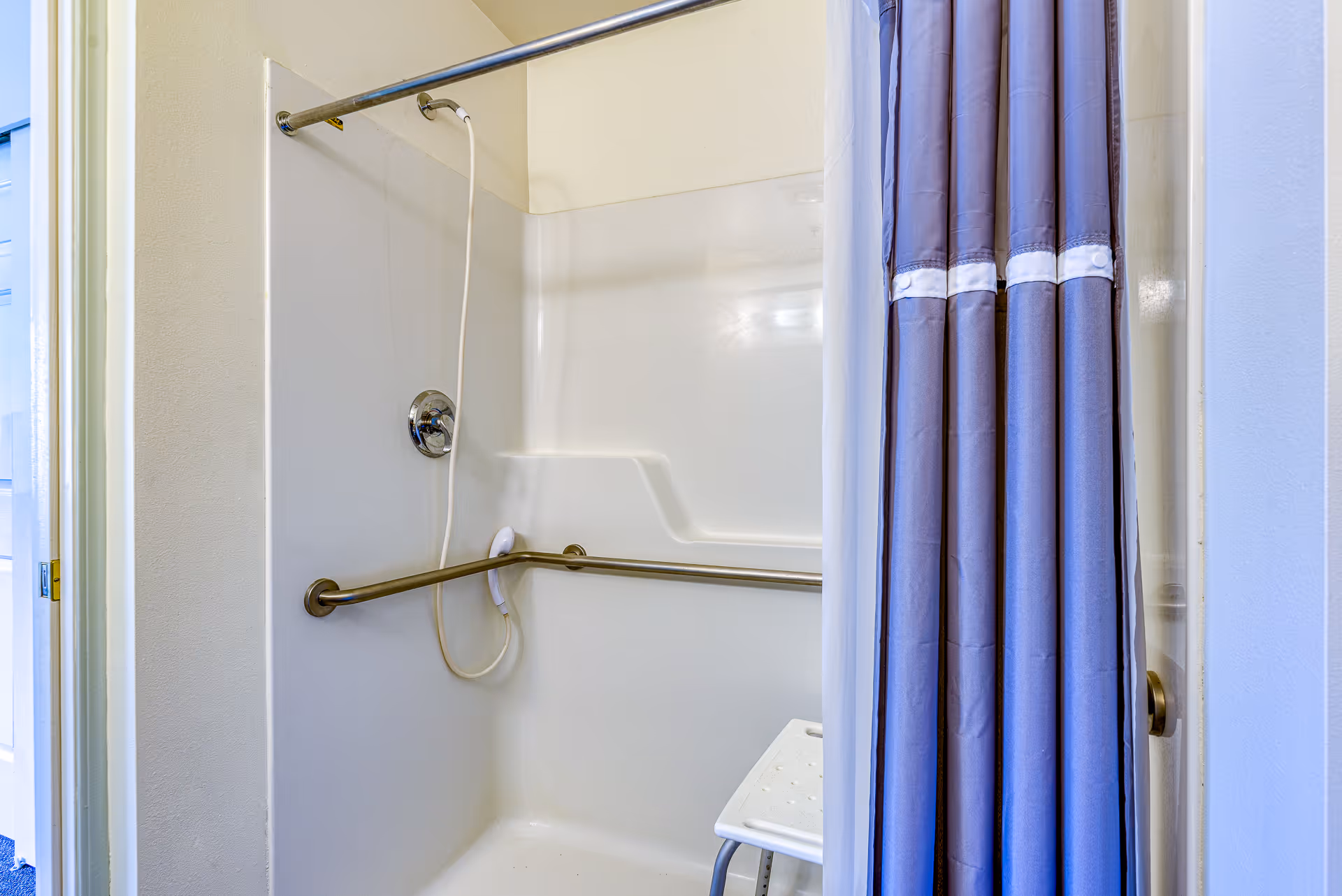 A shower area with a white plastic seat, a handheld showerhead attached to the wall, and a horizontal grab bar. The shower has a white curtain partially drawn to the side and a metal shower curtain rod.