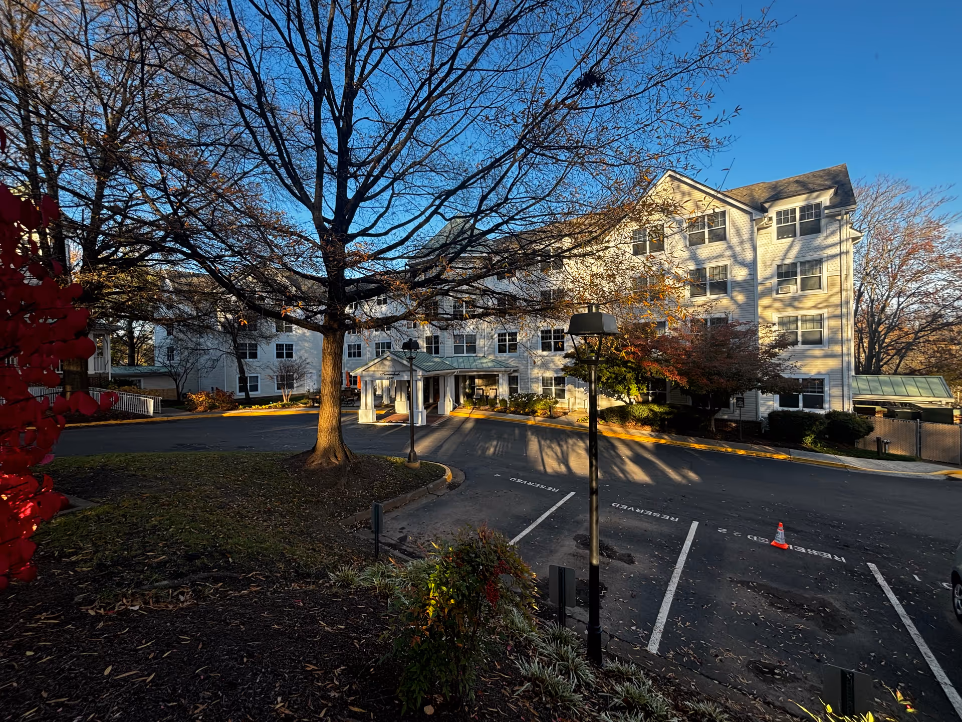 Exterior view of a multi-story white building with many windows, surrounded by trees with autumn foliage. There is a driveway and reserved parking spaces in front of the building, along with a covered entrance. The sky is clear and blue.