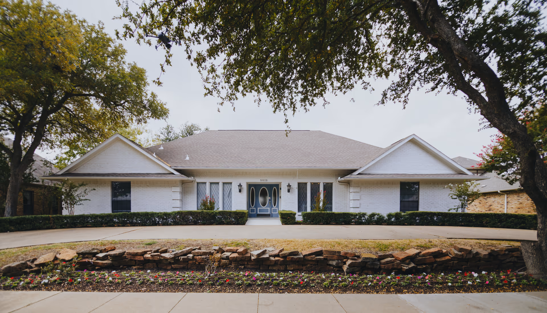Front exterior view of a single-story white brick building with a gray roof, a central entrance featuring double doors with oval windows, flanked by windows with decorative grilles. The building is surrounded by neatly trimmed hedges, a stone-bordered flower bed with colorful flowers, and large trees providing shade.