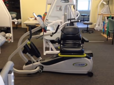 Indoor fitness room with exercise equipment including a NuStep recumbent cross trainer with a black seat, other workout machines, and stools near windows in the background.