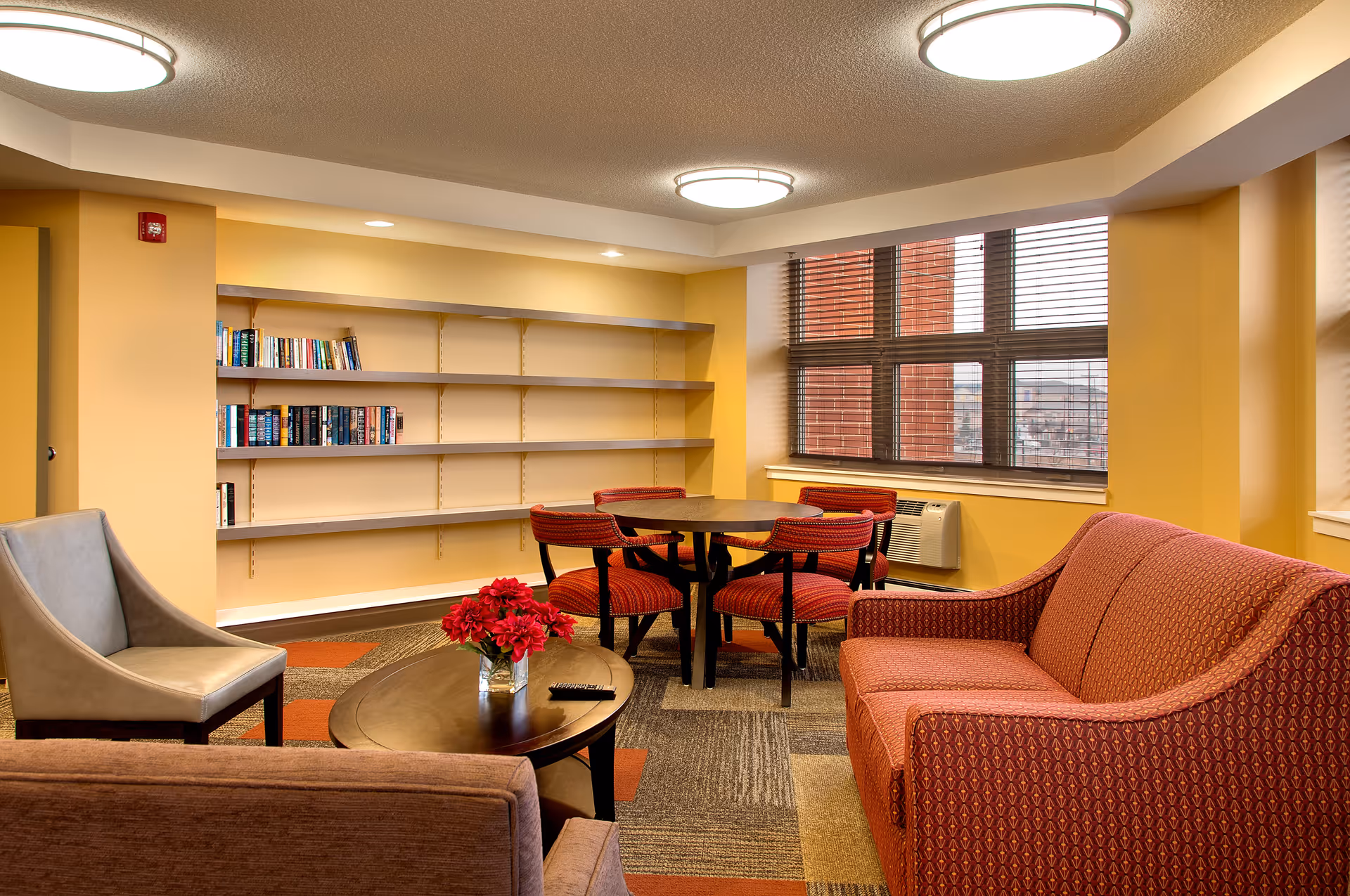 A cozy common area in a senior living facility with a red patterned sofa, a gray armchair, a wooden coffee table with a vase of red flowers, a round table with four red chairs, and a bookshelf with some books. Large windows with blinds allow natural light to enter the room.