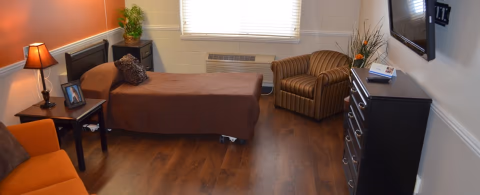 A furnished nursing facility bedroom with a single bed, armchair, dresser, side tables, lamp and wall-mounted TV.
