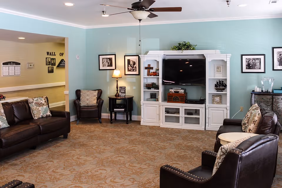 A cozy living room with light blue walls, a ceiling fan, and a patterned carpet. The room features a dark brown leather sofa and two matching armchairs with patterned cushions. A white entertainment center holds a large flat-screen TV and various decorative items. There are framed black and white photos on the walls and a small table with a lamp between the armchairs.