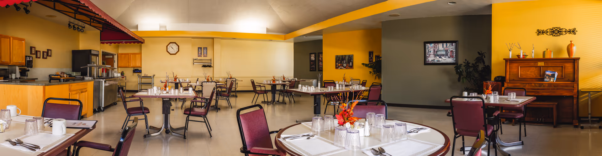 Well-lit dining room with multiple round and rectangular tables set with place settings and chairs, a serving counter to the left and a piano against yellow walls.