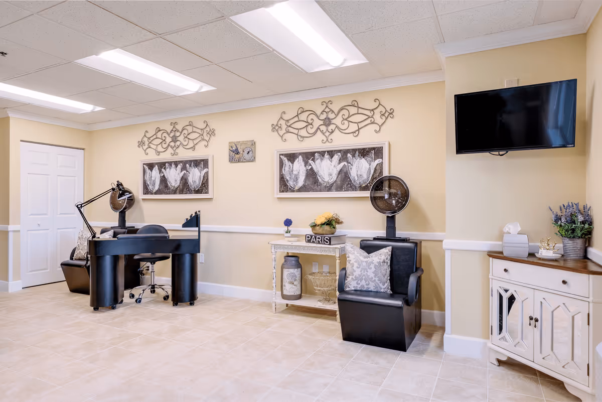 Interior view of a salon area in a senior living facility with two black salon chairs equipped with hair dryers, a small white table decorated with flowers and a Paris sign, wall art featuring white flowers, and a wall-mounted flat screen TV.
