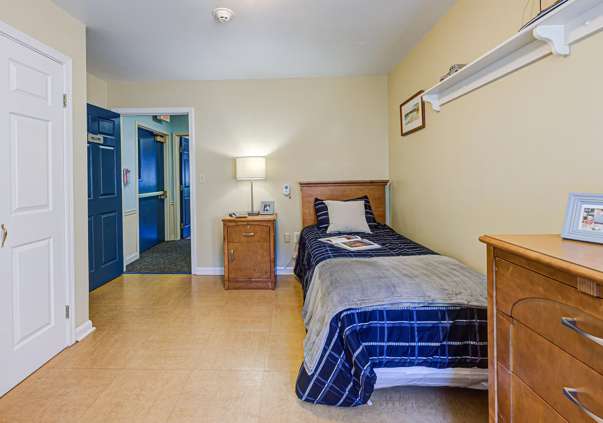 A small, neatly arranged bedroom with a single bed covered in a blue and gray checkered blanket and a gray throw. There is a wooden nightstand with a lamp and a small picture frame next to the bed. A wooden dresser with a framed photo is on the right side of the room. The walls are light beige, and there is a white door on the left side leading to a hallway with blue doors.