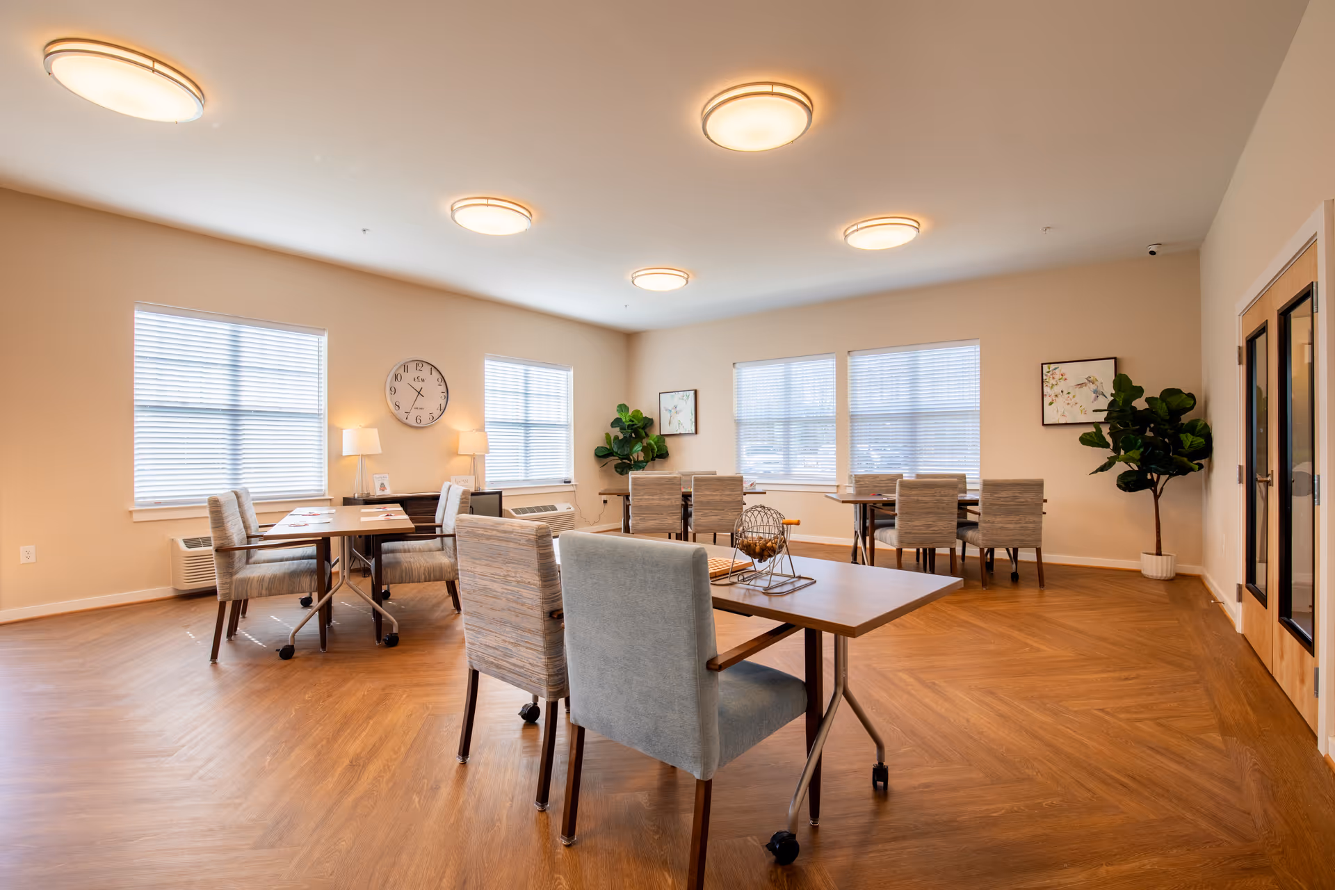 A bright and spacious common room with wooden flooring, several tables and cushioned chairs arranged neatly. The room has large windows with blinds, a wall clock, two table lamps on a sideboard, framed artwork, and potted plants in the corners. The ceiling has multiple round light fixtures providing warm lighting.