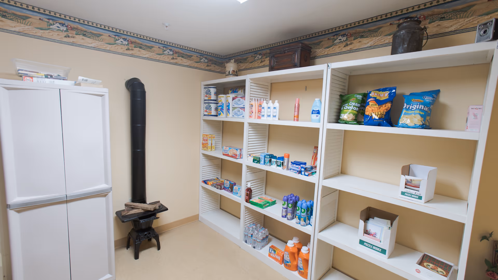 A storage room with white shelves stocked with various household items including snacks, cleaning supplies, bottled water, and toiletries. There is a white refrigerator on the left and a small black wood stove with logs on it. The walls are beige with a decorative border near the ceiling featuring a countryside scene.