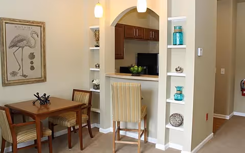 Interior view of a dining area with a wooden table and three chairs, adjacent to a kitchen pass-through window with a high chair. The walls are light-colored with built-in shelves displaying decorative items, and a framed flamingo artwork is hung on the wall.