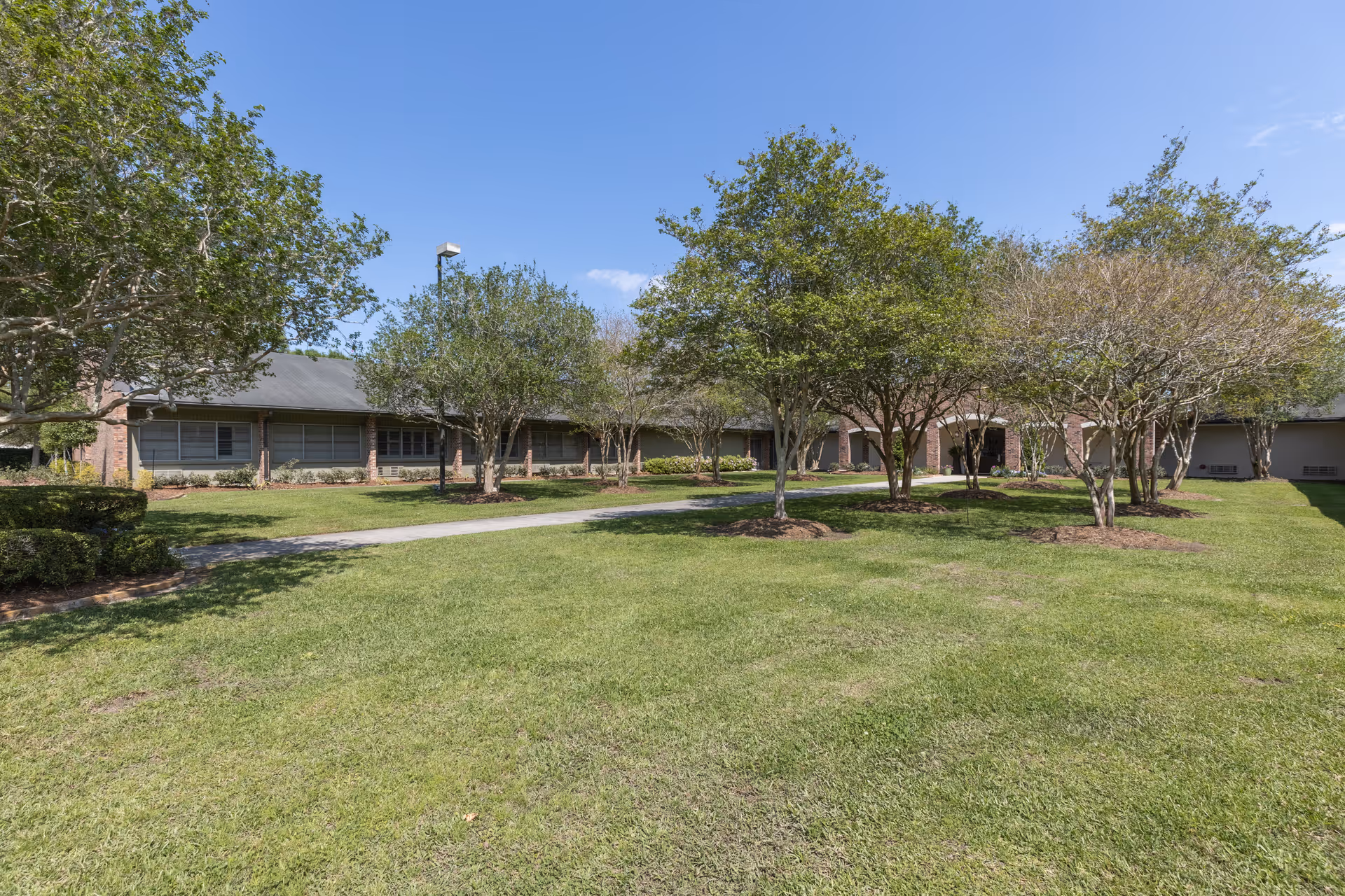 A single-story brick building with multiple windows surrounded by a well-maintained grassy lawn and several trees under a clear blue sky.