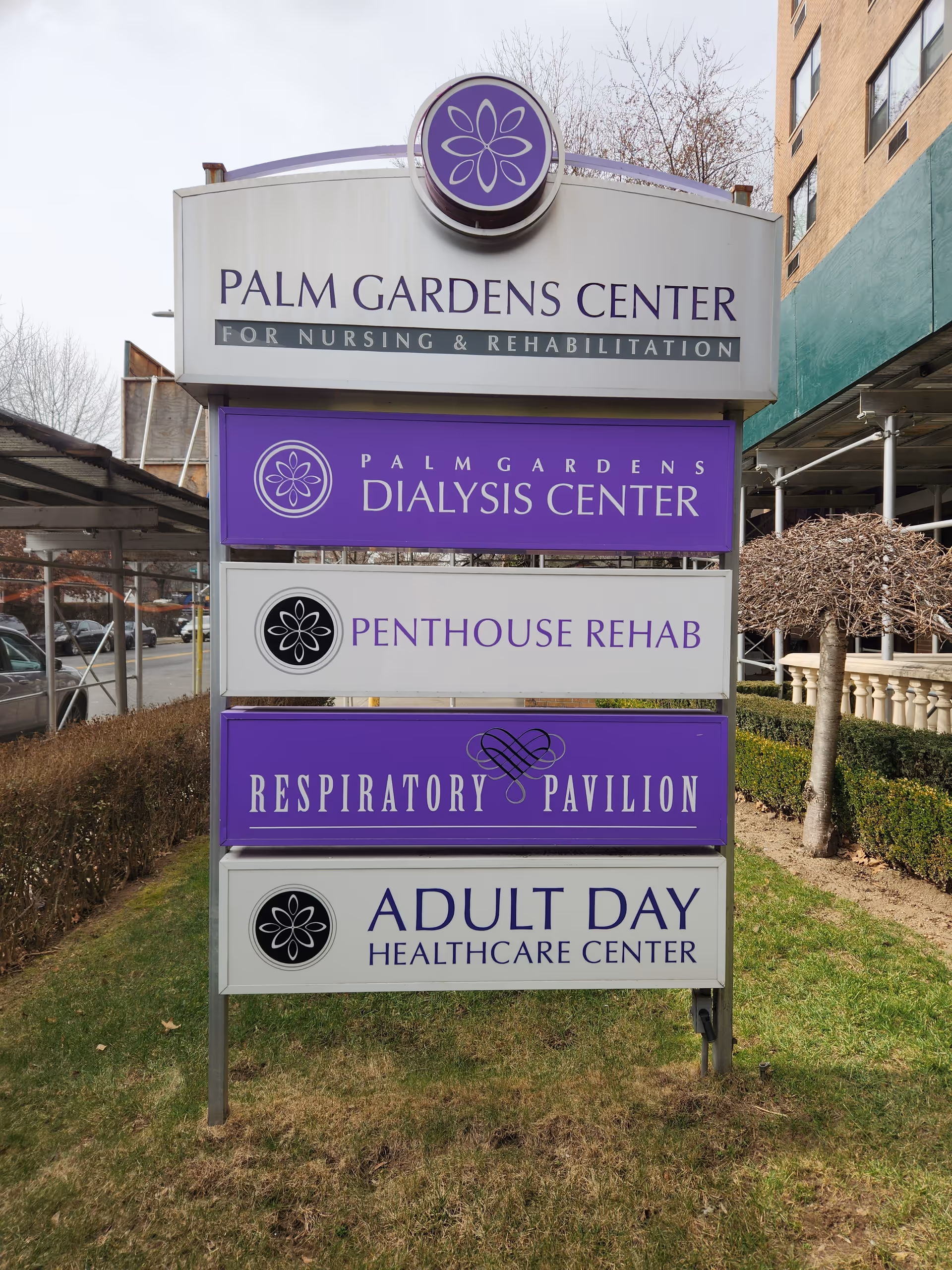 Outdoor sign for Palm Gardens Center for Nursing & Rehabilitation with sections for Dialysis Center, Penthouse Rehab, Respiratory Pavilion, and Adult Day Healthcare Center, situated on a grassy area near a sidewalk and buildings.