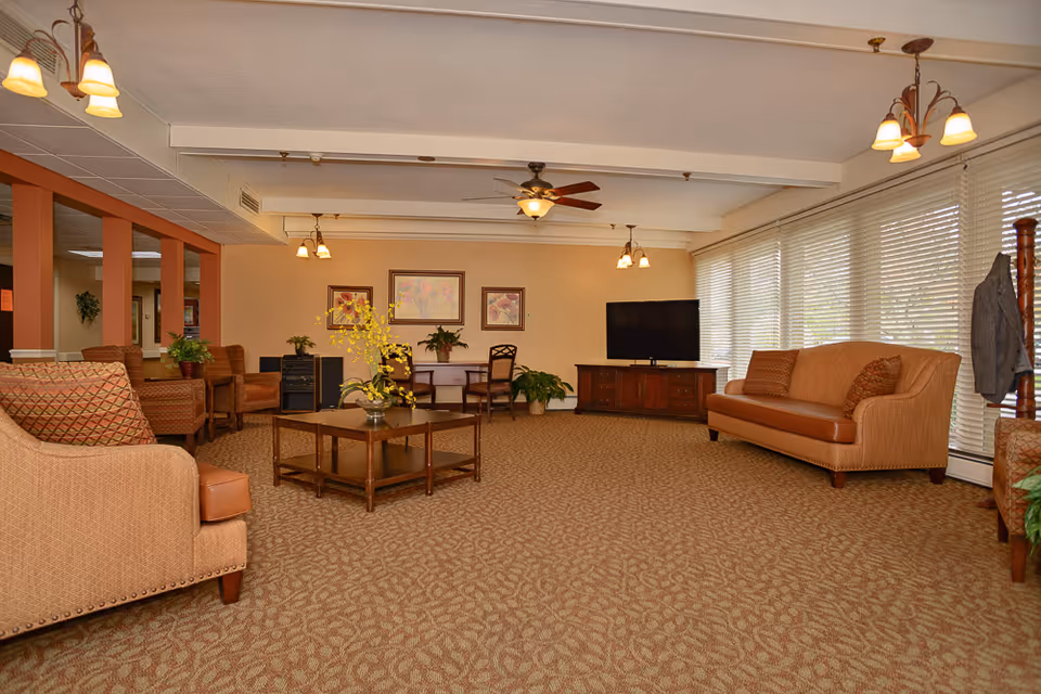 Spacious senior living common room with sofas, coffee table, TV and large windows with blinds.