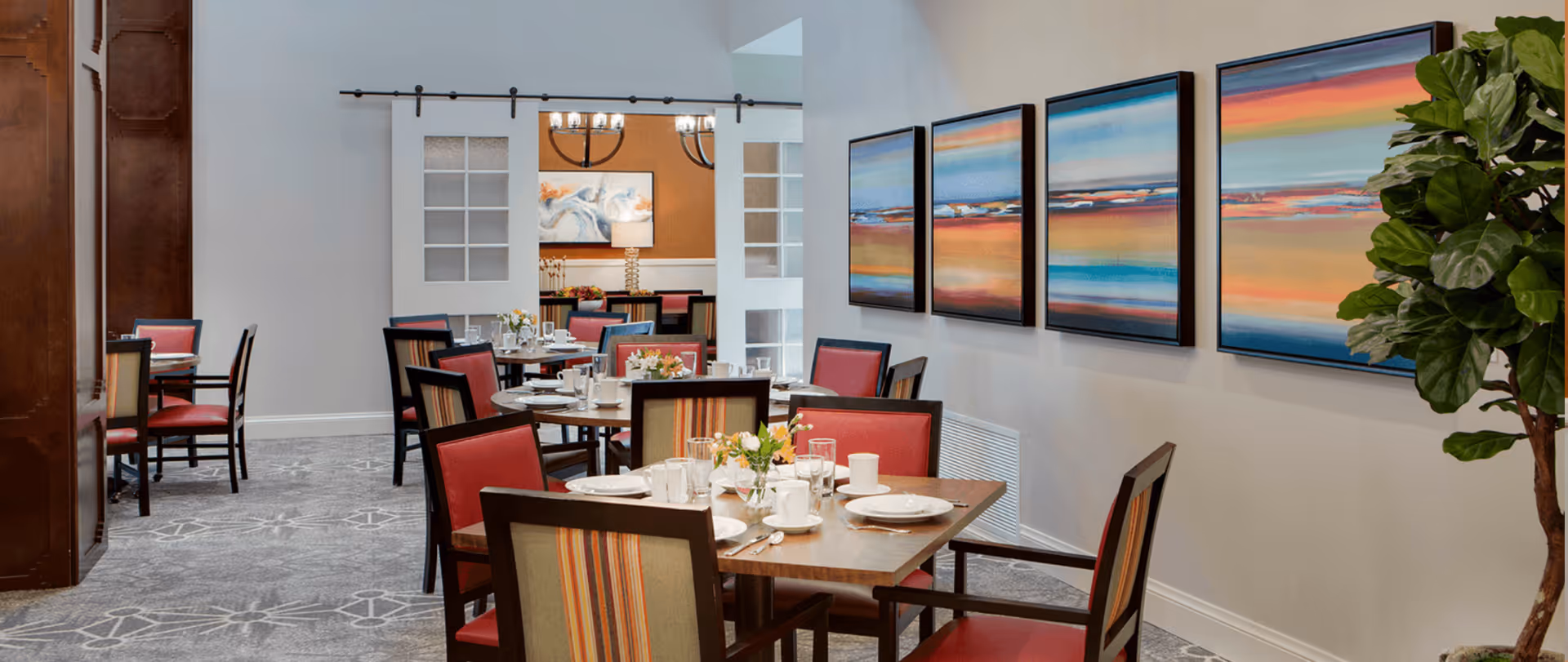 A dining room with several wooden tables set with white plates, cups, and glassware. The chairs have red cushions and striped backs. The room features a patterned carpet, three colorful landscape paintings on the wall, and a large green plant in the corner. In the background, there is another dining area visible through a wide doorway with sliding glass doors.