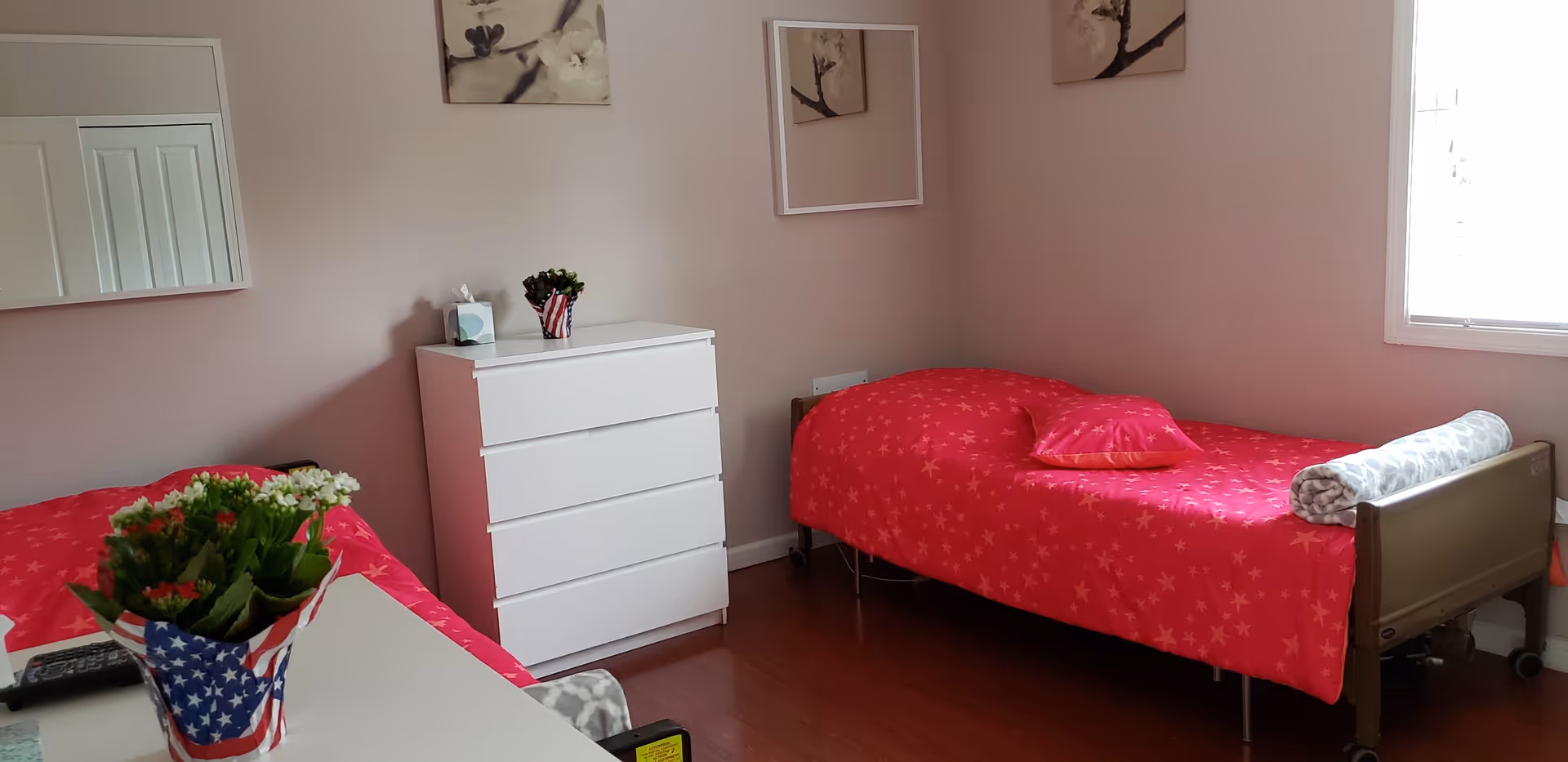 A small bedroom with two single beds covered in red star-patterned bedspreads. One bed has a rolled blanket at the foot and a matching pillow. There is a white chest of drawers between the beds with a small plant and a tissue box on top. The walls are light-colored with framed artwork and a mirror. A window on the right side lets in natural light.