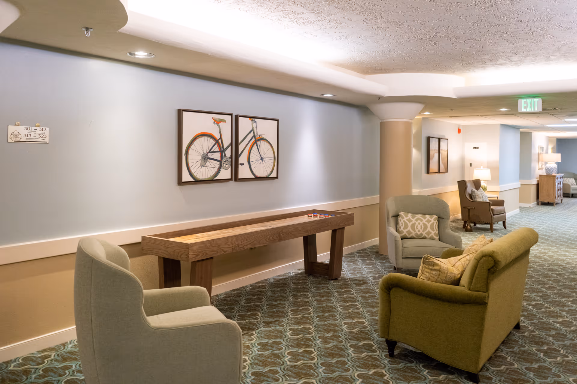 A cozy hallway seating area in a senior living facility featuring several upholstered chairs and a loveseat arranged around a wooden shuffleboard table. The walls are painted light blue and beige, decorated with framed artwork of a bicycle. The carpet has a patterned design, and there are lamps and exit signs visible in the background.