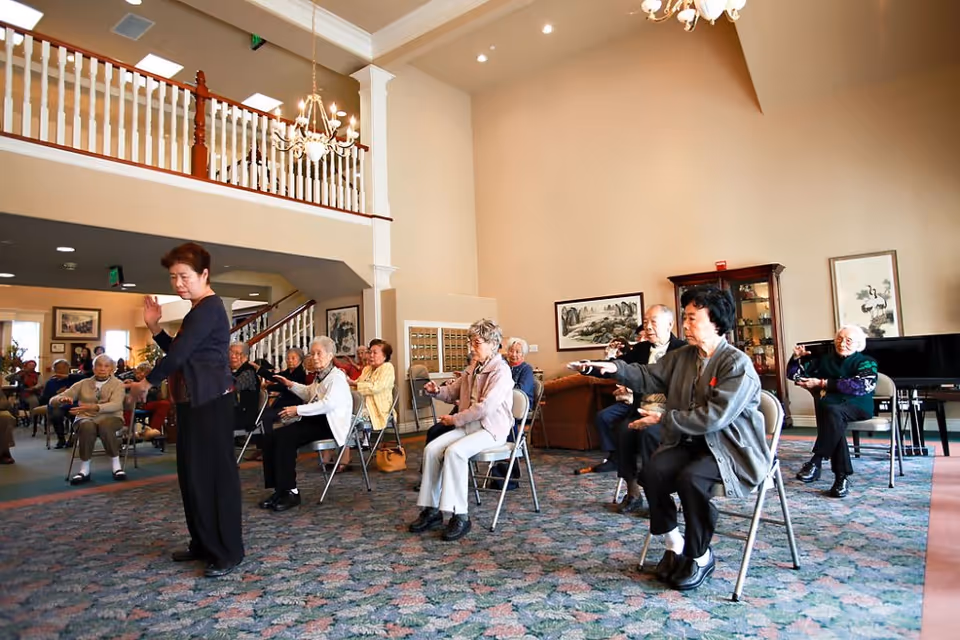 A group of elderly individuals seated on chairs in a spacious, well-lit room participating in a seated exercise or Tai Chi session led by an instructor standing in front of them. The room has high ceilings, chandeliers, framed artwork on the walls, and a carpeted floor.