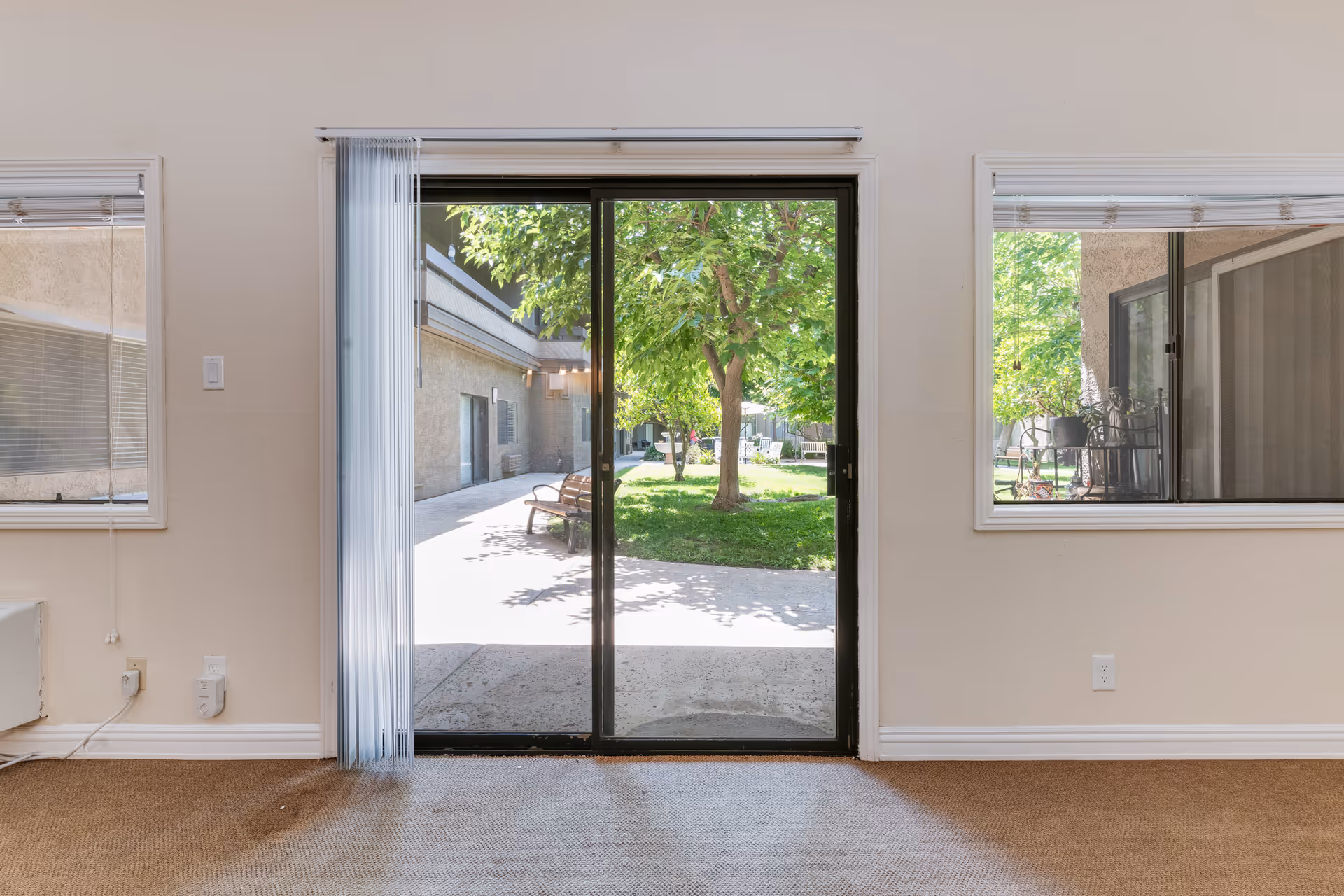View from inside a room looking out through a glass sliding door to a courtyard with green grass, trees, benches, and surrounding building walls. The room has beige walls, carpeted floor, and two windows with blinds on either side of the sliding door.