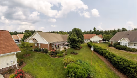 Aerial view of single-story cottage-style homes around a landscaped green courtyard with trees and a cloudy sky.