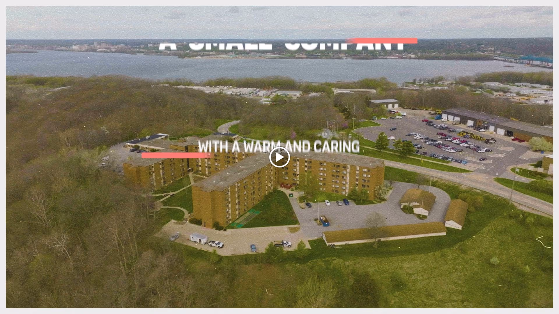 Aerial view of a large multi-story brick building surrounded by trees and parking lots, with a river and bridge visible in the background. The image includes partially visible text overlay that reads 'A SMALL COMPANY WITH A WARM AND CARING'.