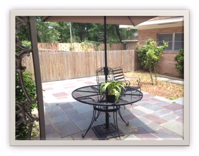 Outdoor patio area with a round black metal table and matching chairs. A potted plant is placed on the table under a large umbrella. The patio is paved with square tiles and is enclosed by a wooden fence. There are some small trees and bushes near a brick building in the background.