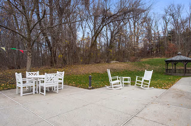 Outdoor patio area with white wooden chairs and tables on a concrete surface, surrounded by grass and leafless trees, with a gazebo visible in the background.