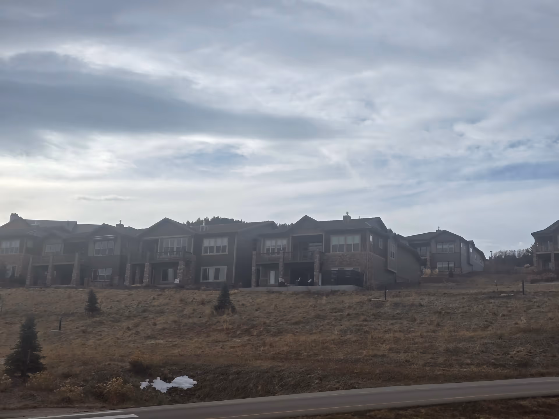 A row of residential buildings on a hillside under a cloudy sky, with sparse small trees and dry grass in the foreground.